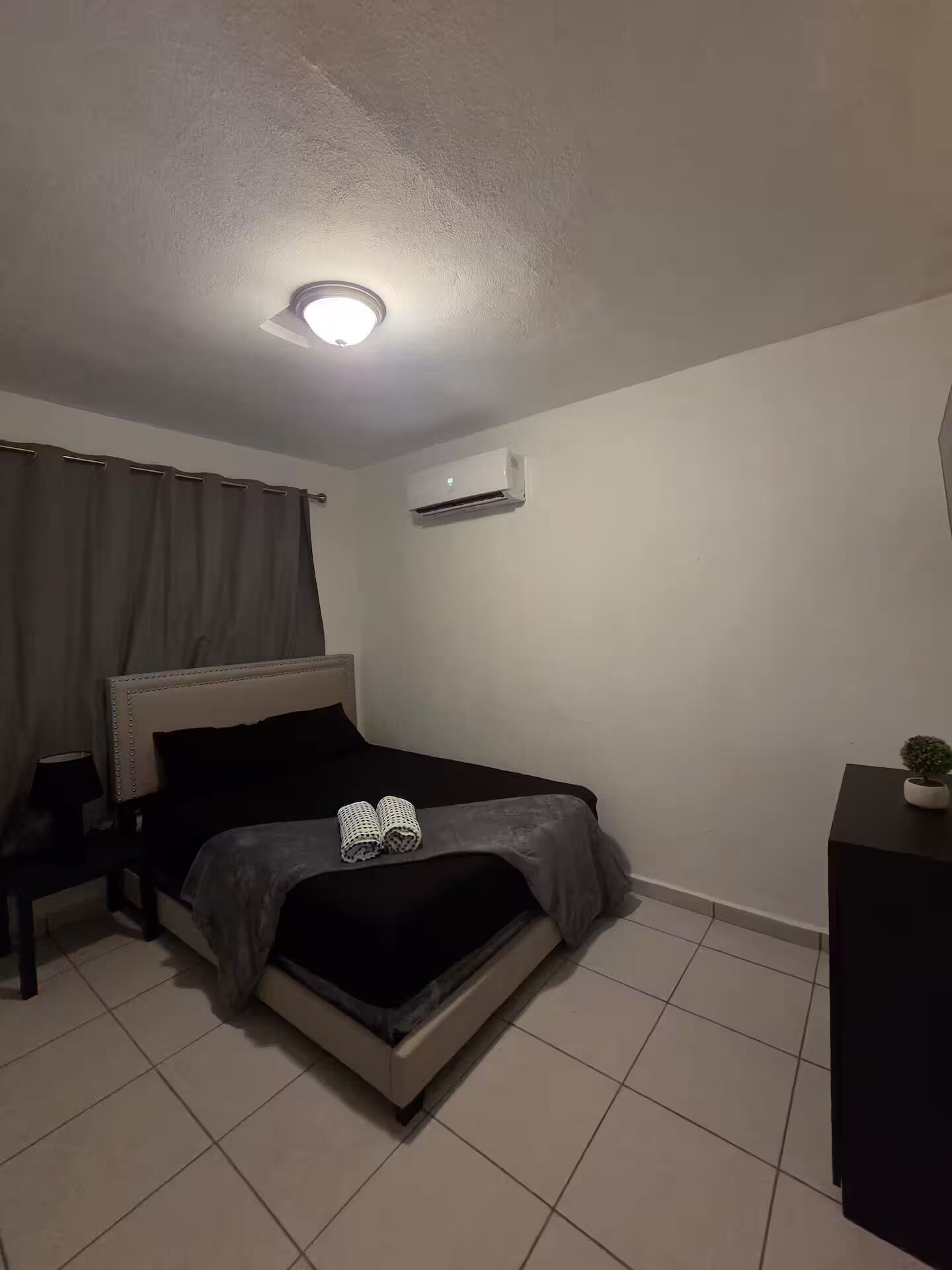 Une chambre à coucher comprenant un lit, une table de chevet, une commode, un appareil de climatisation et un luminaire au plafond.