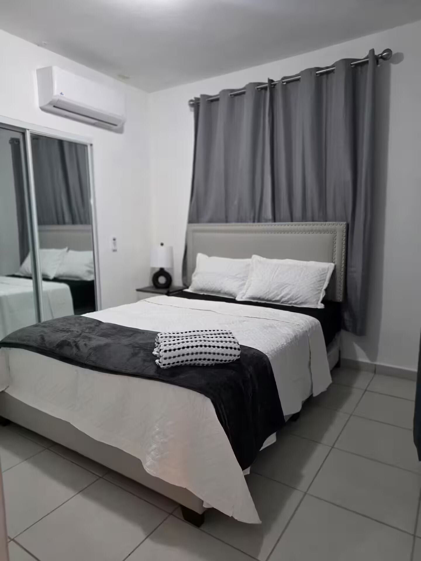 Een slaapkamer met een bed, nachtkastje, lamp en raam met gordijnen.