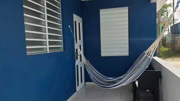 Una habitación con paredes azules, una puerta blanca, una ventana con persianas horizontales, una hamaca y un piso de baldosas.