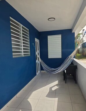 Una habitación con paredes azules, una puerta blanca, una ventana con persianas horizontales, una hamaca y un piso de baldosas.