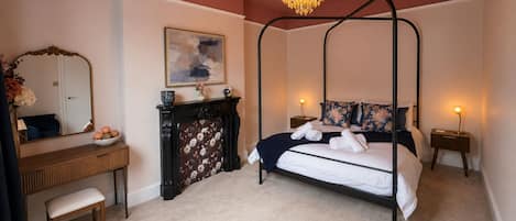 A four-poster bed with floral bedding, a mirror, a vase with flowers, and a fireplace with a floral-patterned screen.
