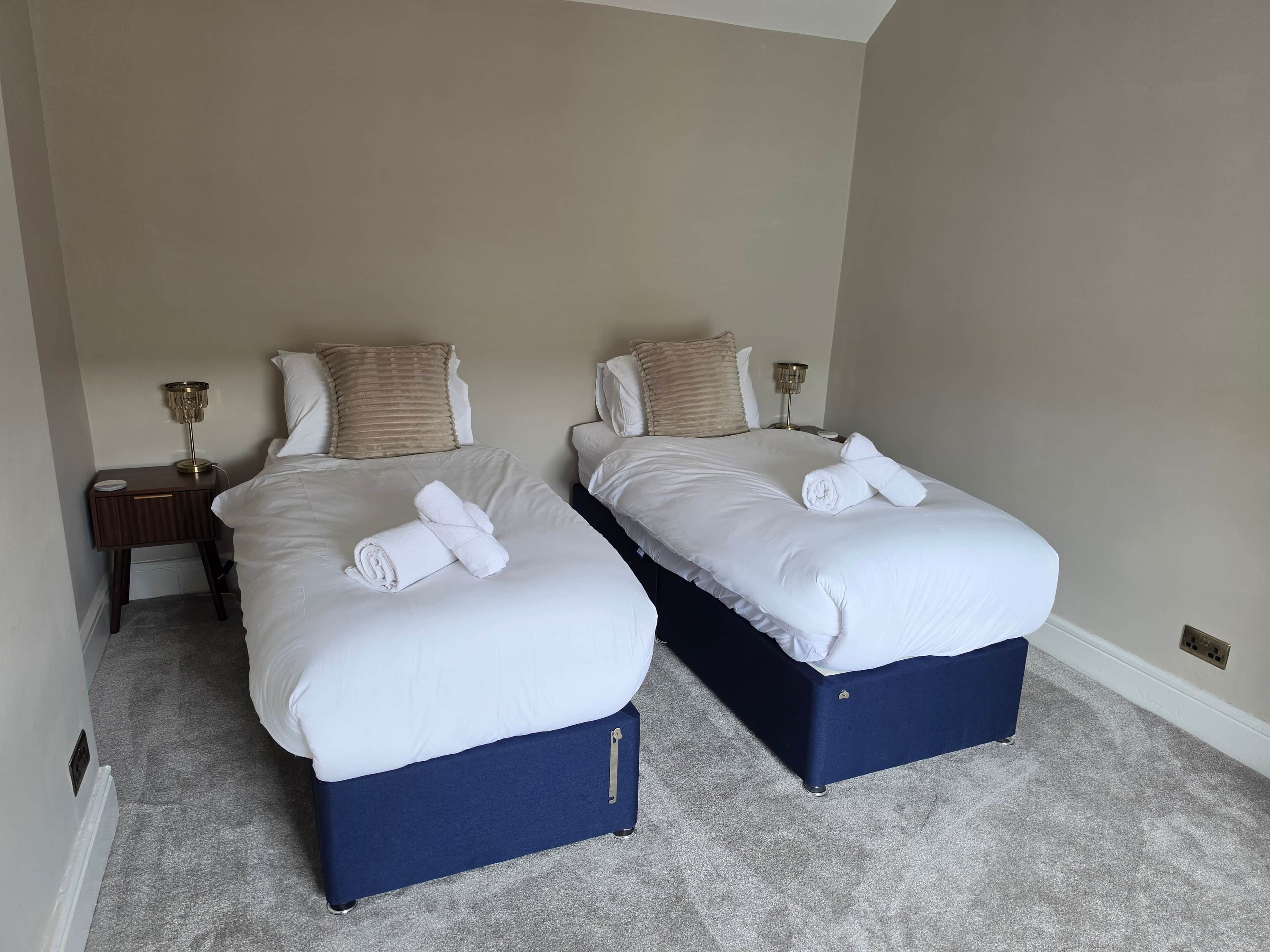 Twee enkele bedden met witte lakens en blauwe bedpoten, een nachtkastje met een lamp, en een vloerbedekking.