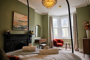 Une chambre avec une cheminée, un fauteuil rouge, un lit avec du linge de maison blanc, un tableau et une grande fenêtre avec des rideaux.