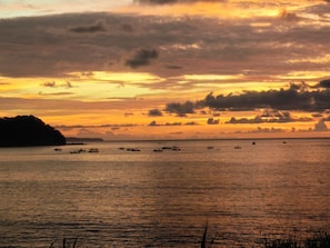 Un coucher de soleil sur une mer calme, avec des bateaux, une terre lointaine et un ciel nuageux.