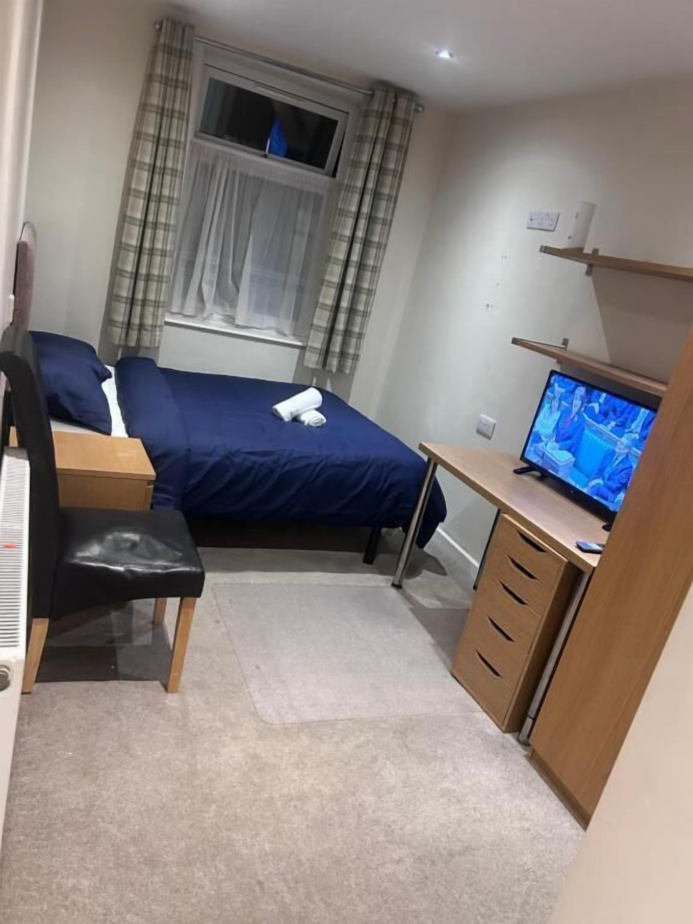 A bedroom with a bed, a desk with a computer, a chair, a window with curtains, and a television.