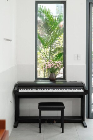 A piano with a bench in front, a window with a view of greenery and pink flowers, and a power outlet on the wall. - Green Apartment near My Khe Beach and Dragon Bridge -R302 (Đà Nẵng)