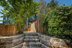A set of stone steps leading up to a wooden staircase, surrounded by greenery and a wooden fence.