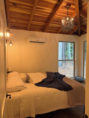 Un dormitorio con una cama grande, un candelabro, un equipo de aire acondicionado y una ventana con vista a árboles.
