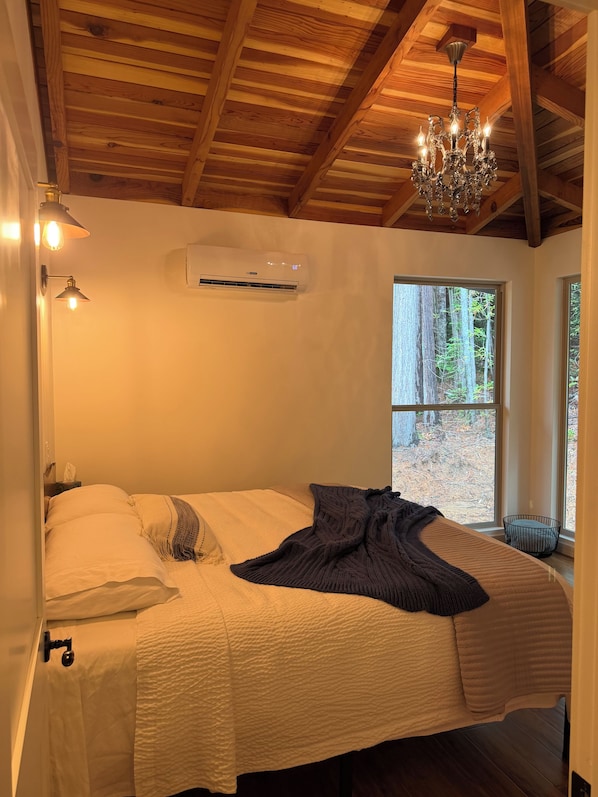 A bedroom with a large bed, a chandelier, an air conditioning unit, and a window with a view of trees. - Brand New custom built 2 bedroom home in Hiouchi near Stout Grove/Titan Trail (Hiouchi)