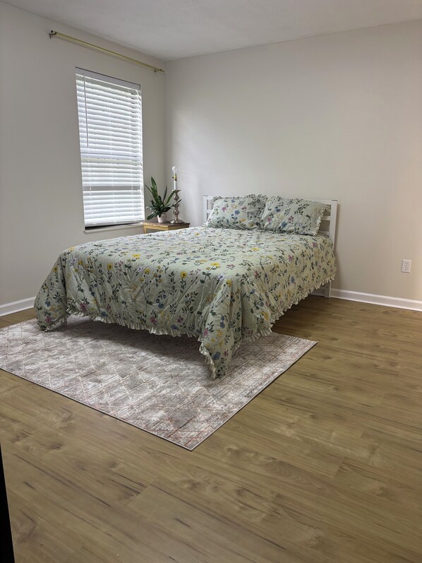 A bedroom with a bed, a window with blinds, a bedside table with a lamp, and a potted plant.