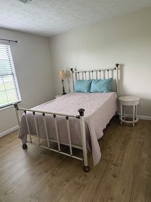 A bedroom with a metal-framed bed, a bedside table, a lamp, and a window with blinds.