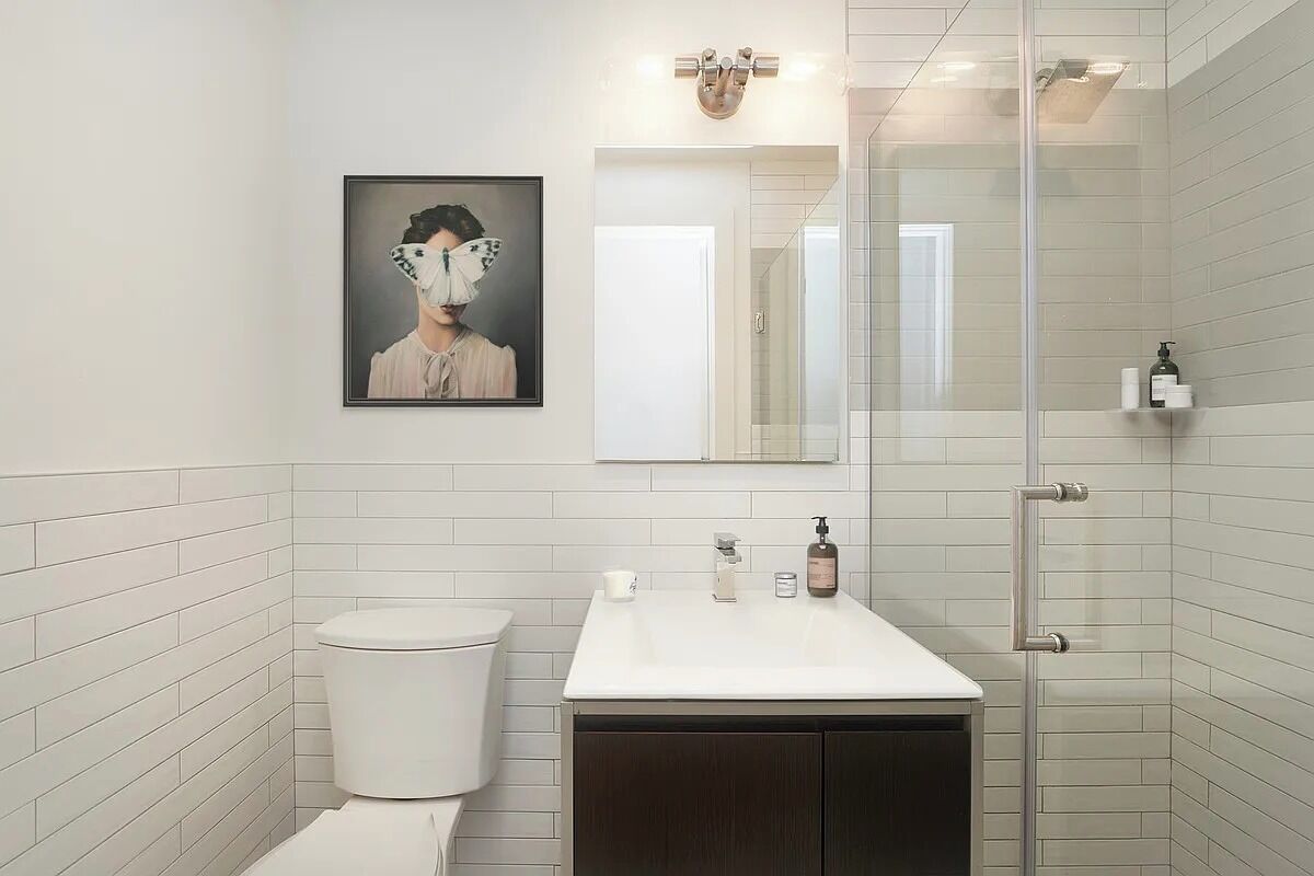Een badkamer met witte betegelde wanden, een wit wastafelmeubel, een donker kastje, een glazen douchecabine, een kunstwerk in een lijst aan de muur en een spiegel boven de wastafel.