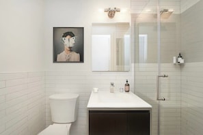 A bathroom with white tiled walls, a white sink, a dark cabinet, a glass shower enclosure, a framed artwork on the wall, and a mirror above the sink.