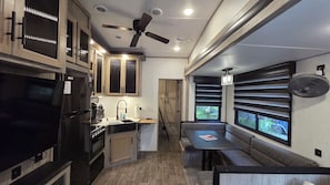 A compact kitchen with a stove, sink, and cabinets, a dining area with a table and bench, and a window with blinds.
