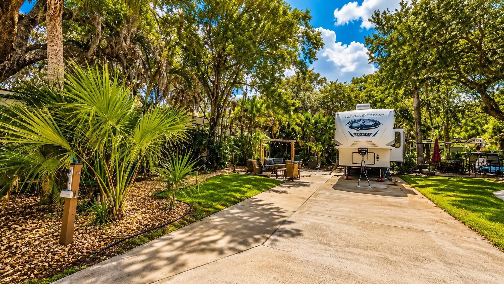 A recreational vehicle (RV) with a 'RIVERESTORY' logo parked in a landscaped area with a concrete path, surrounded by trees and greenery.