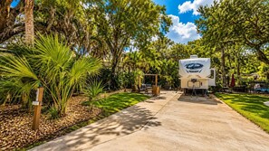 A recreational vehicle (RV) with a 'RIVERESTORY' logo parked in a landscaped area with a concrete path, surrounded by trees and greenery. - Happy Hour Hideaway RV Rental at River Ranch! 247 (River Ranch)