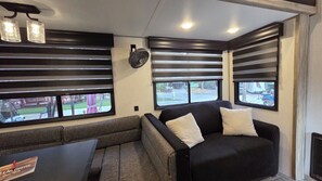 A compact RV interior with a sofa, two windows, a fan, and a table. - Happy Hour Hideaway RV Rental at River Ranch! 247 (River Ranch)