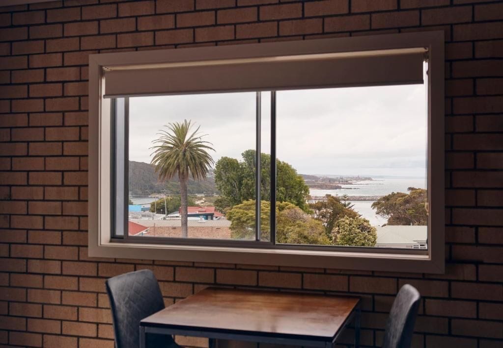 Una habitación con pared de ladrillo, ventana con vista al paisaje costero y una mesa con dos sillas.