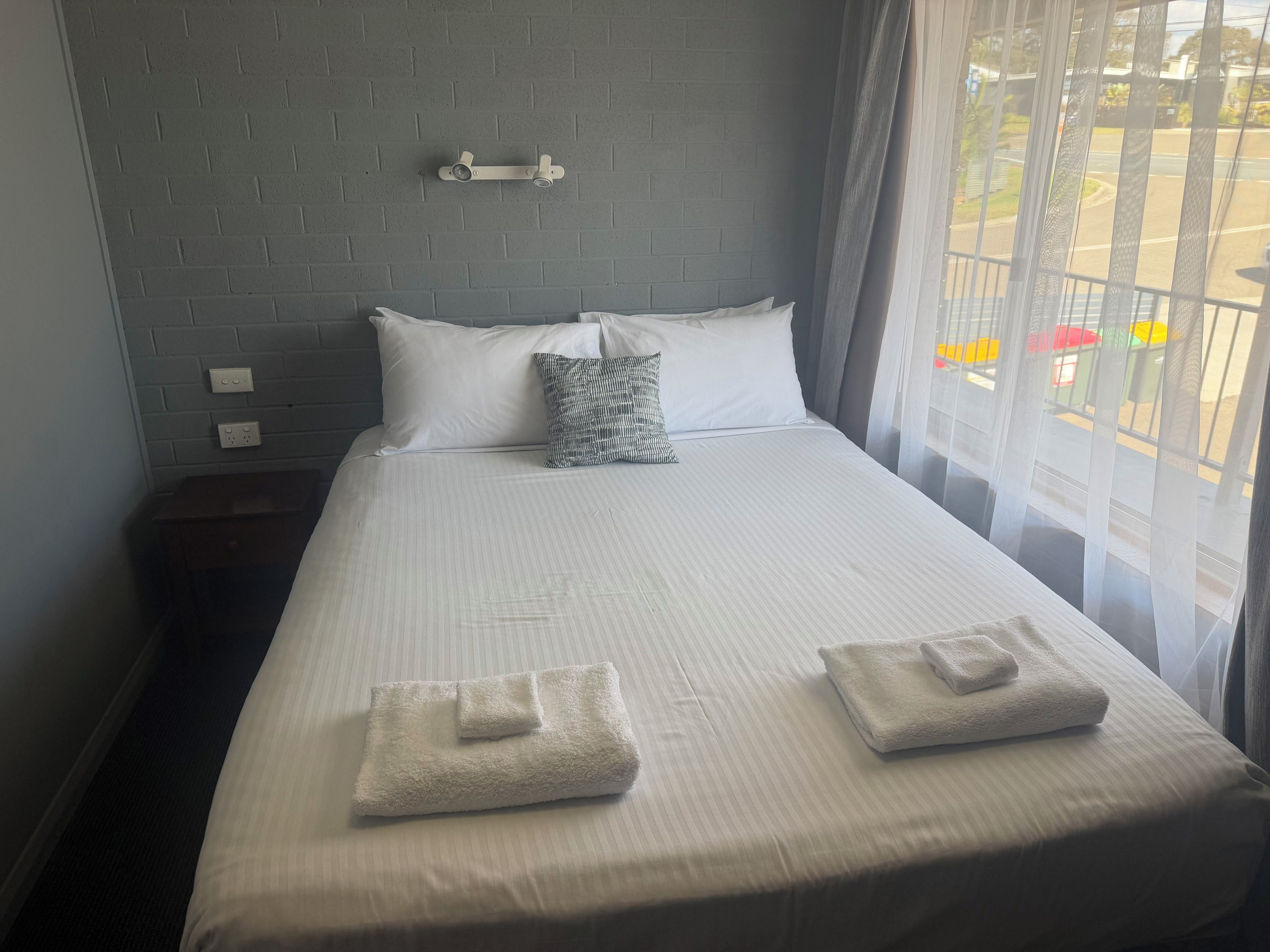 A neatly made bed with white linens and two folded towels, a gray pillow, and a wall-mounted light fixture.