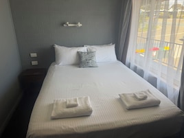 Cama bem arrumada com lençóis brancos e duas toalhas dobradas, um travesseiro cinza e um aplique de parede.