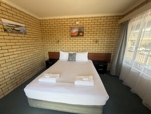 A hotel room with a brick wall, a bed with white linens, two bedside tables, and a painting above the headboard. - Coastal Comfort Motel (Narooma)
