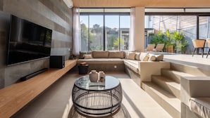 Una sala de estar moderna con un televisor de pantalla plana, un sofá modular, una mesa de centro y una escalera que lleva a un balcón con mobiliario exterior.