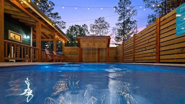 Piscina com deck de madeira e espreguiçadeiras, cercada por um muro de madeira e luzes de corda.