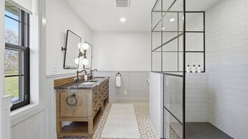 A bathroom with a wooden vanity, a mirror with lights, a shower with glass doors, and patterned floor tiles.
