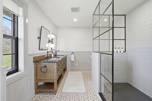 A bathroom with a wooden vanity, a mirror with lights, a shower with glass doors, and patterned floor tiles. - Welcome to The Sunset Lakehouse, a private waterfront retreat where modern farmhouse design meets the natural beauty of Prince Edward County. (Stinson Block Rd, Consecon, ON K0K 1T0, Canada)