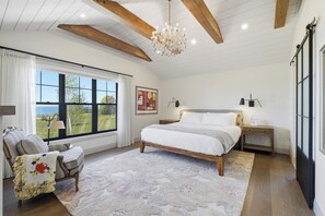 A bedroom with a large bed, a chandelier, a window with a view, and a chair with a floral cushion. - Welcome to The Sunset Lakehouse, a private waterfront retreat where modern farmhouse design meets the natural beauty of Prince Edward County. (Stinson Block Rd, Consecon, ON K0K 1T0, Canada)