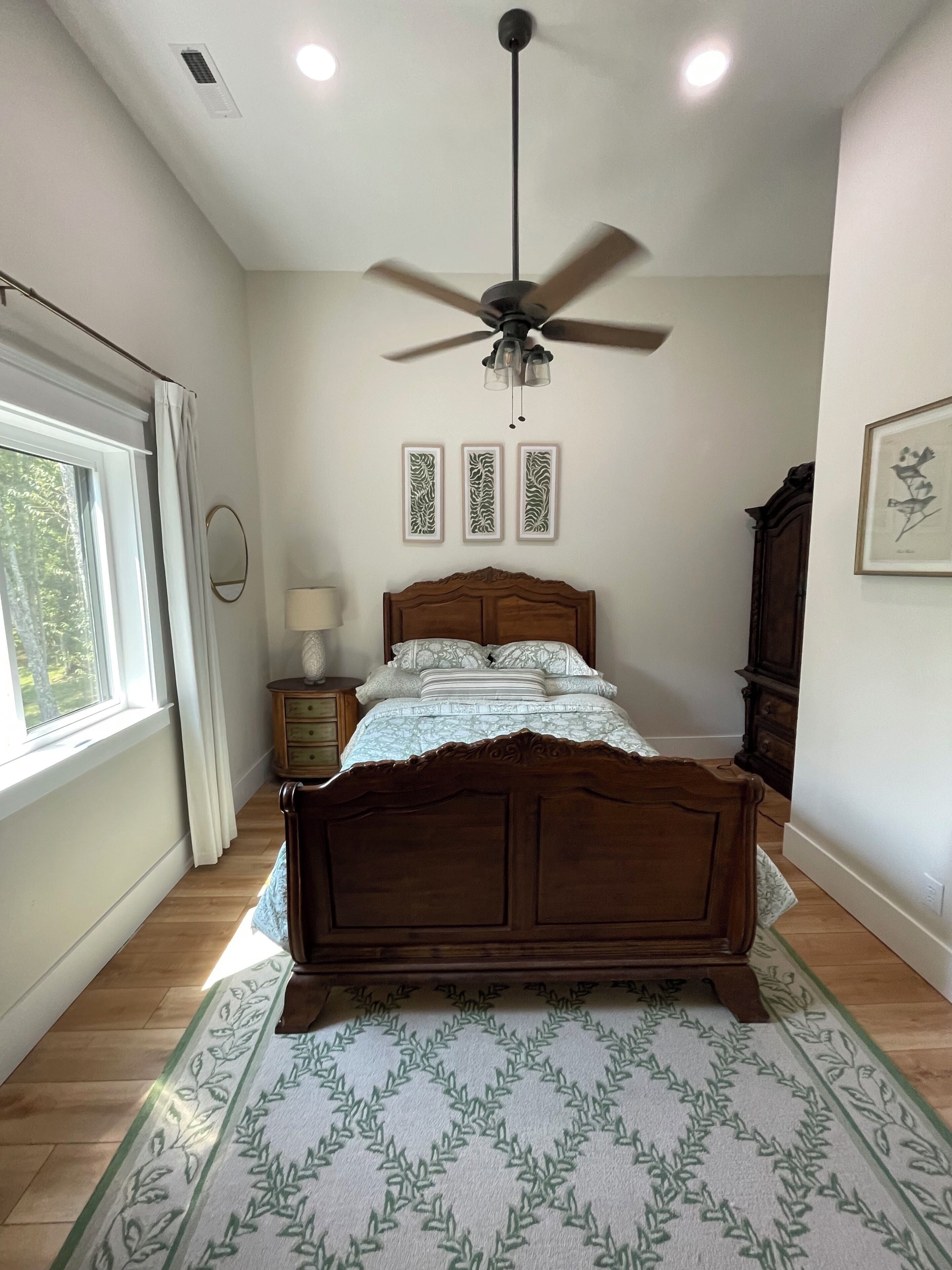 A bedroom with a wooden bed, a ceiling fan, a window with curtains, and framed artwork on the wall.