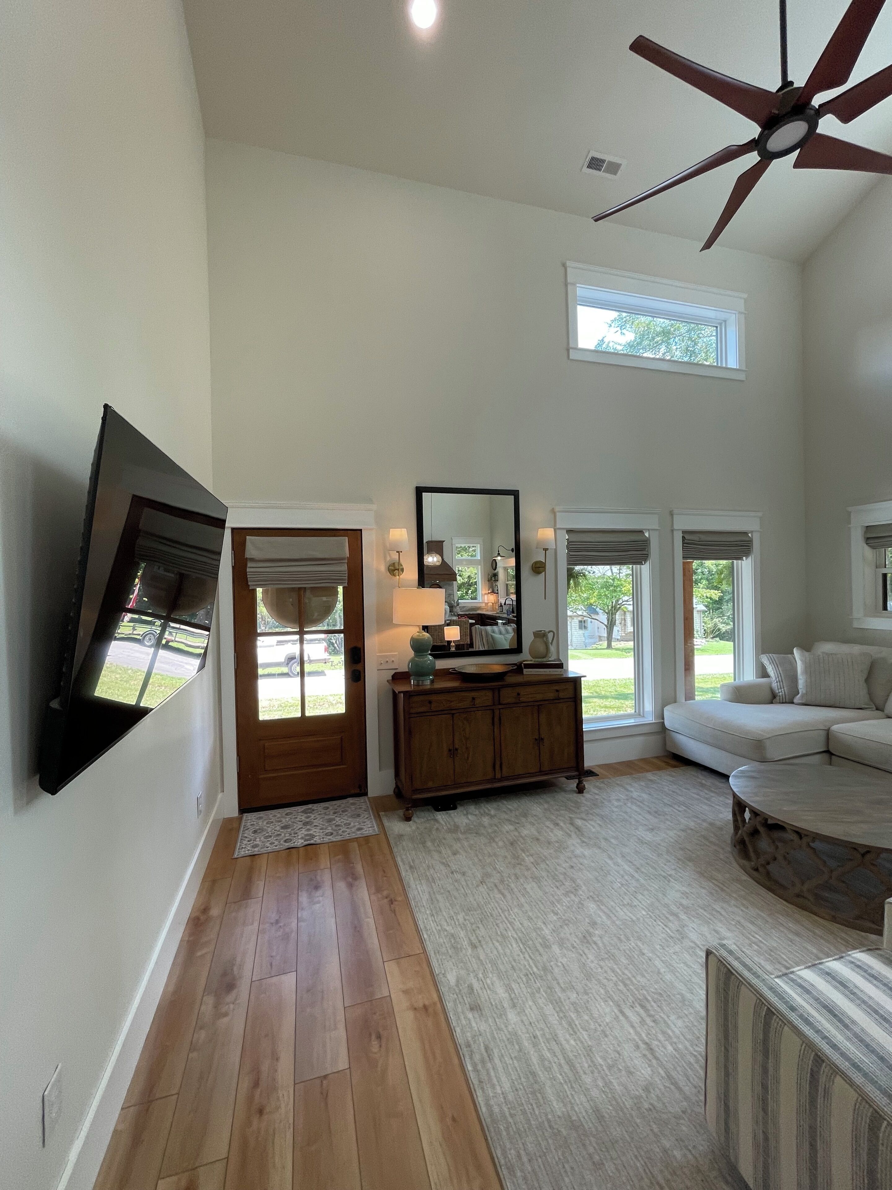 A spacious living room with a large ceiling fan, a flat-screen TV mounted on the wall, a wooden cabinet, and a sectional sofa.