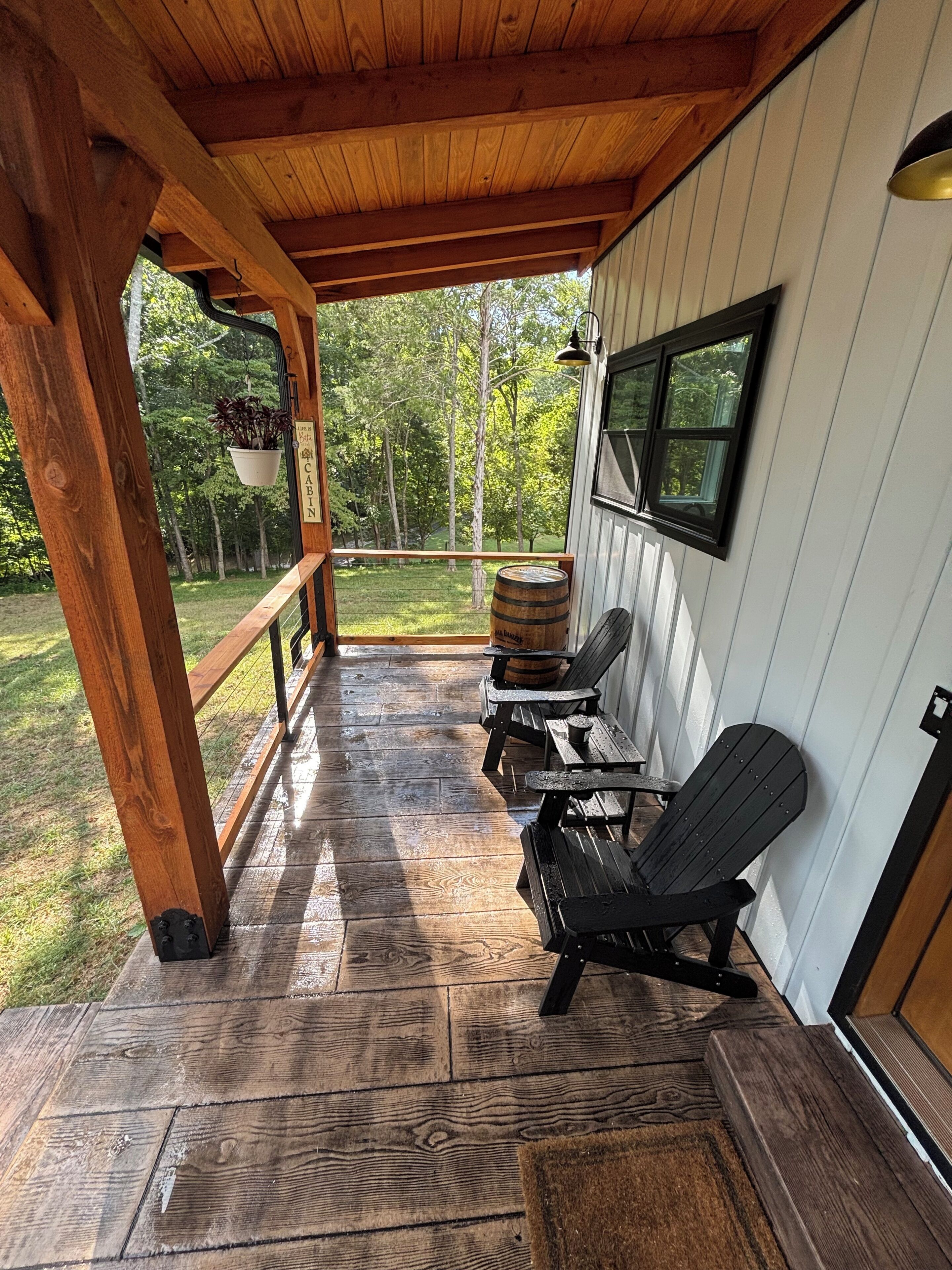 A covered porch with wooden flooring, black Adirondack chairs, a barrel, and a hanging plant.
