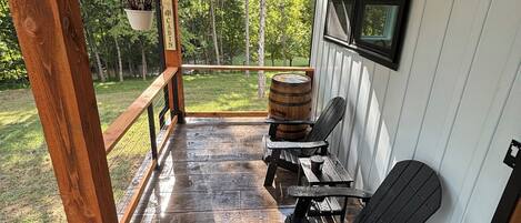 A covered porch with wooden flooring, black Adirondack chairs, a barrel, and a hanging plant.