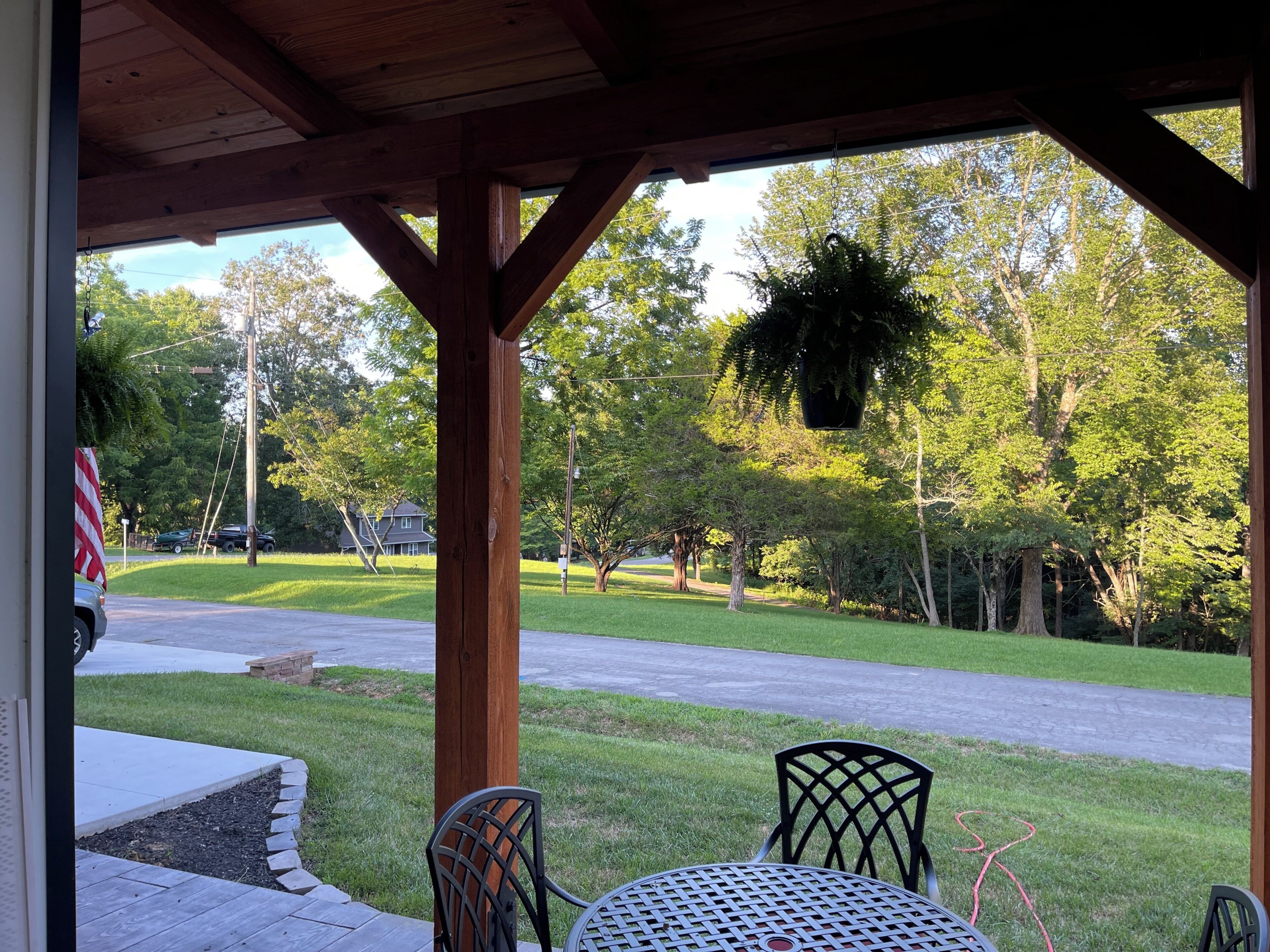 A covered patio with a table and chairs, a hanging planter, and a view of a residential street and greenery.