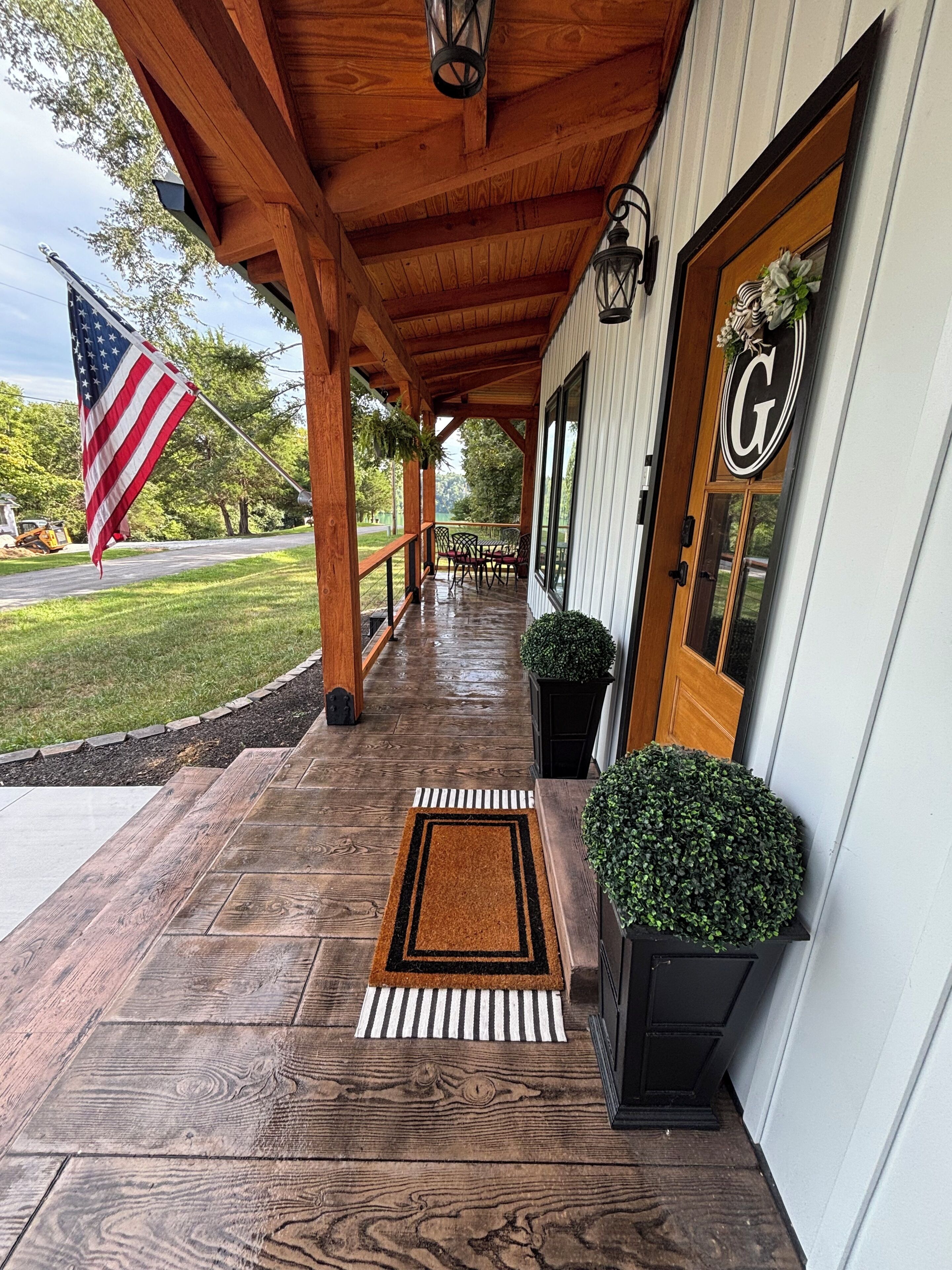 A wooden covered porch with a doormat, potted plants, and an American flag.