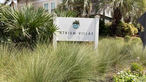 En skylt för Atrium Villas med en logotyp, omgiven av högt gräs och palmer.