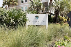 A sign for Atrium Villas with a logo, surrounded by tall grass and palm trees. - Oyster Shell Villa by Sandpiper | Near Beach (Seabrook Island)