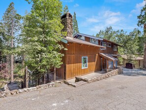 Une cabane en bois à deux étages, dotée d’une cheminée en pierre, entourée d’arbres et bordée d’une allée en gravier.