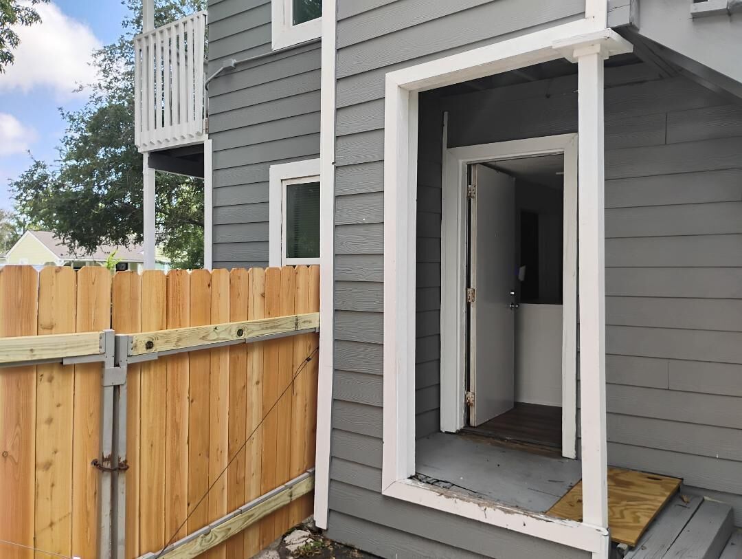 A residential building with a grey exterior, a wooden fence, and a door leading to an interior space.