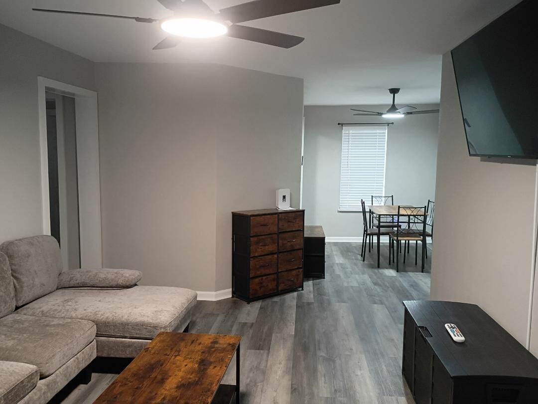 A living room with a sofa, a coffee table, a TV stand, a ceiling fan, and a dining area with a table and chairs.