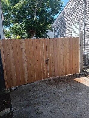 Een houten hek met een metalen poort, een oprit van beton en een huis met een stenen gevel.