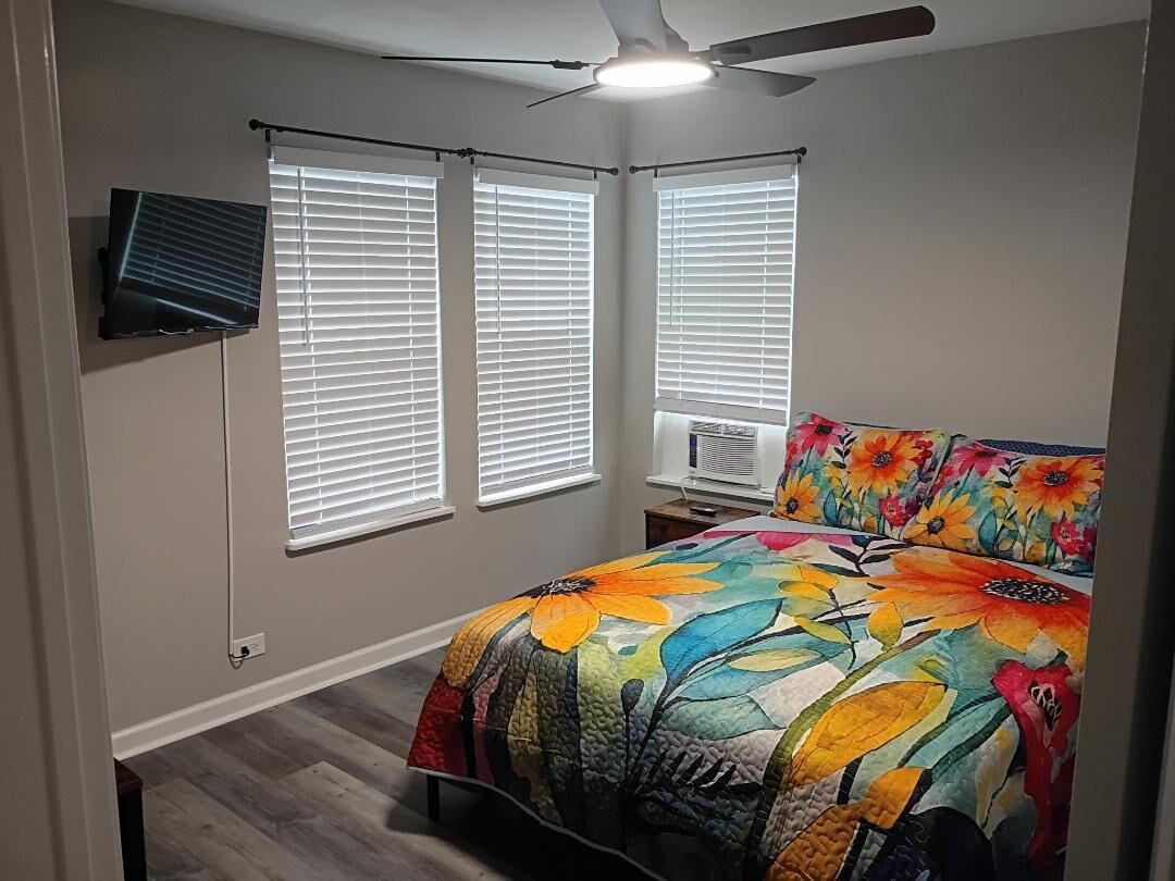 A bedroom with a bed, two windows with blinds, a ceiling fan, and a wall-mounted TV.