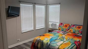 A bedroom with a bed, two windows with blinds, a ceiling fan, and a wall-mounted TV.