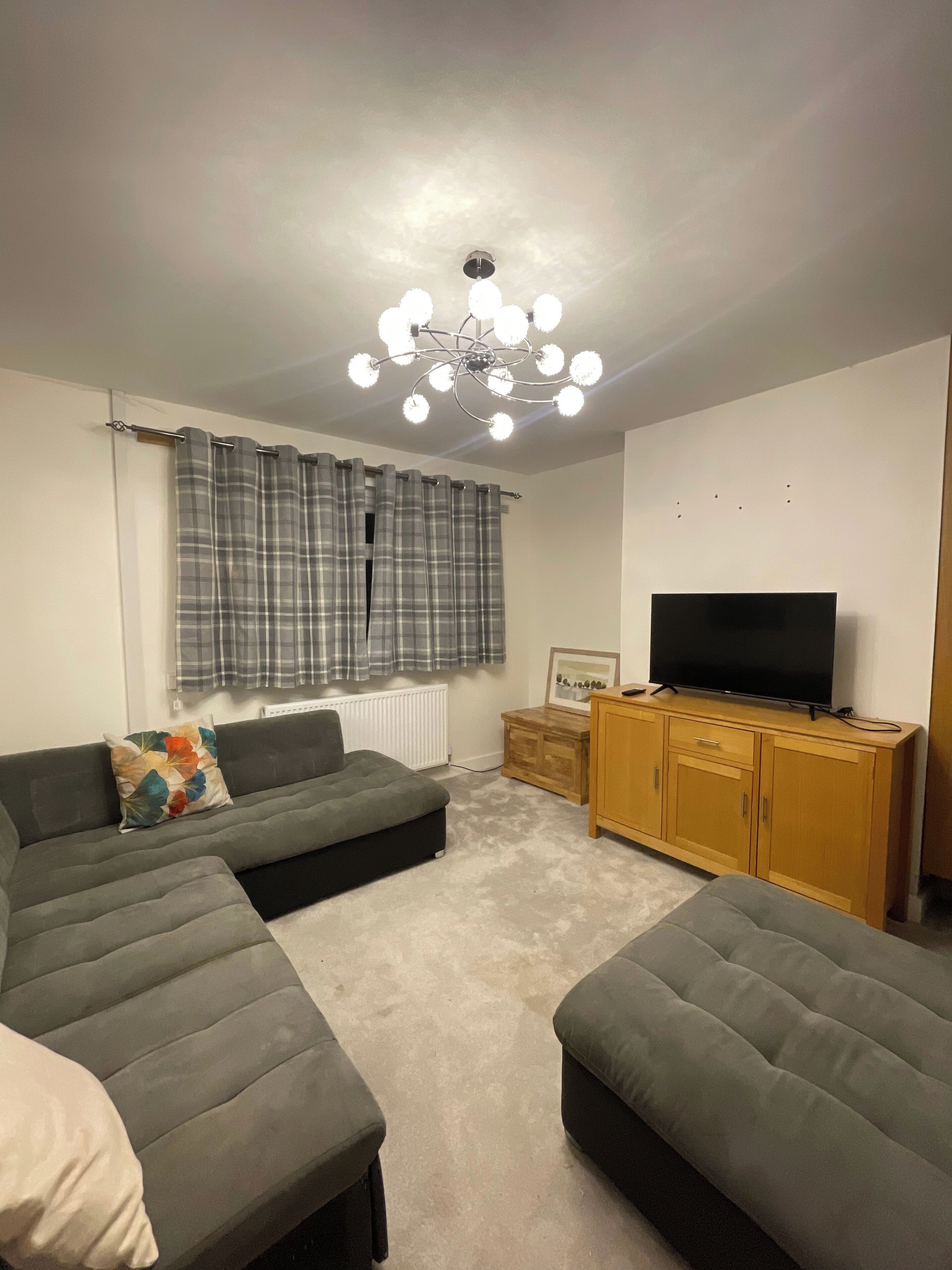Un salón con un sofá modular, reposapiés, un televisor sobre un mueble de madera, una araña y cortinas de cuadros.