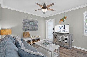 Ein Wohnzimmer mit grauem Sofa, einem weißen Couchtisch, einem Deckenventilator und einem an der Wand montierten Fernseher, der 'AM ANN MARIA RENTAL PROPERTIES' anzeigt.