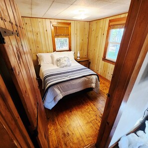 Ein Schlafzimmer mit Holzboden, einem Bett mit gestreifter Bettdecke, zwei Fenstern mit Jalousien und einem Nachttisch mit Lampe.