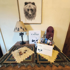 Ein Willkommensschild, ein gerahmtes Bärenbild, eine Lampe und ein Tisch mit einer Patchworkdecke und Informationsbroschüren.