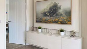 A hallway with a white console table, a painting of a tree and landscape, and a door leading to another room. - 3-Bed House, Sleeps 7 with Parking & Garden (Wishaw)