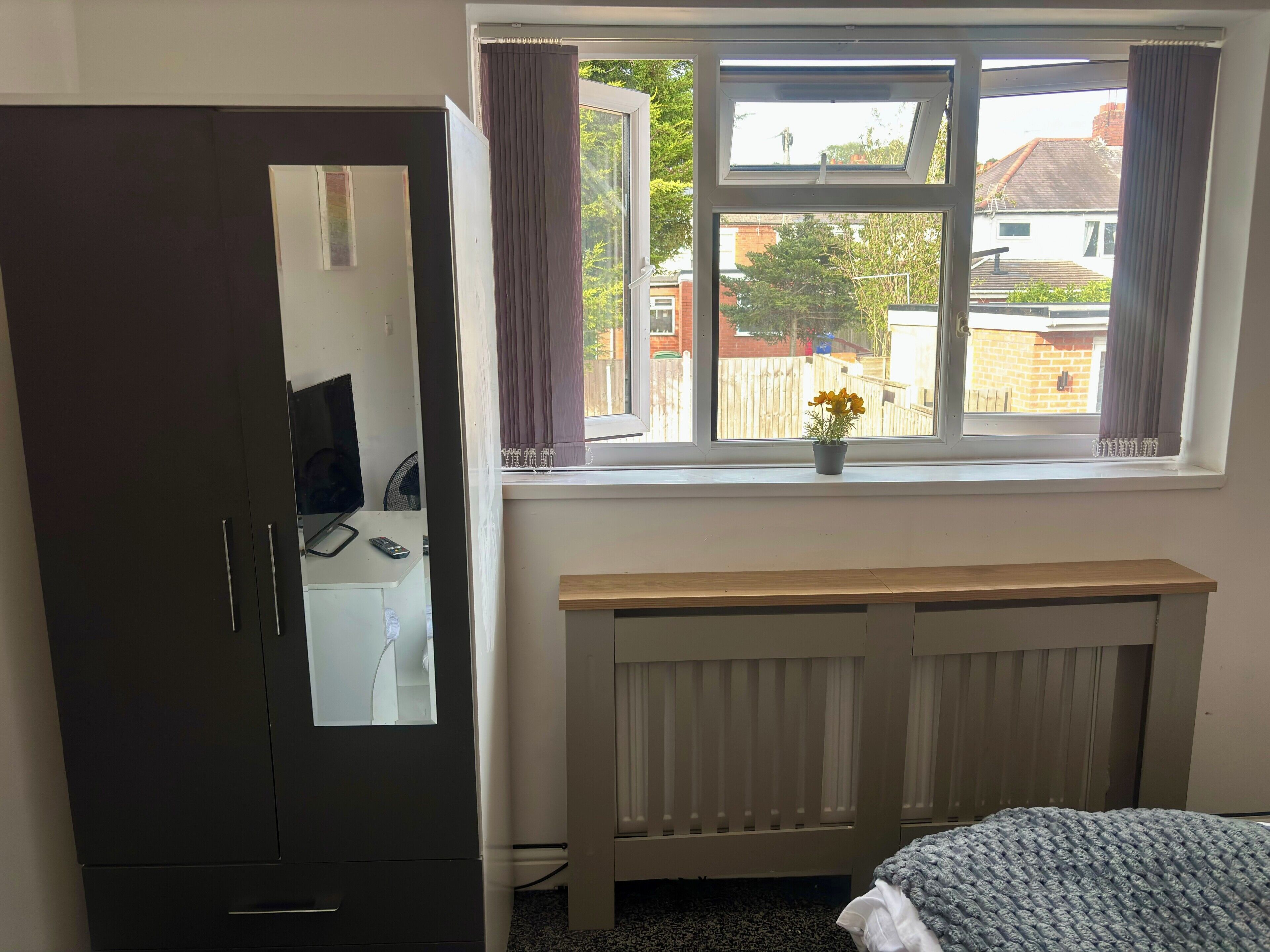 Ein Schlafzimmer mit Bett, Schrank, Heizkörper, einem Fenster mit Vorhängen und Blick nach draußen.